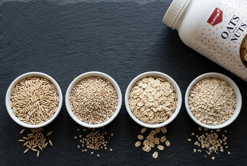 Four types of oats side by side in white bowls on dark slate, groats, steel-cut, rolled and quick oats, with a Sunnah Grains Oats with Nuts jar at the edge