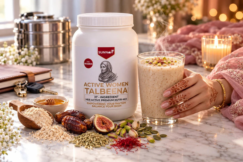 A woman's hennaed hand holding a glass of creamy Active Women Talbeena beside the Sunnah Grains Active Women Talbeena jar on white marble, with saffron, fennel, dates, figs, pistachios and pumpkin seeds