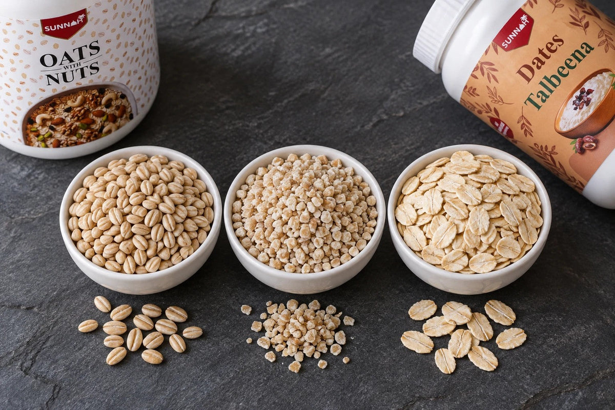 Whole barley (jau), cracked barley (jau ka dalia) and rolled oats (jai) in three white bowls on slate, with Sunnah Grains Dates Talbeena and Oats with Nuts jars